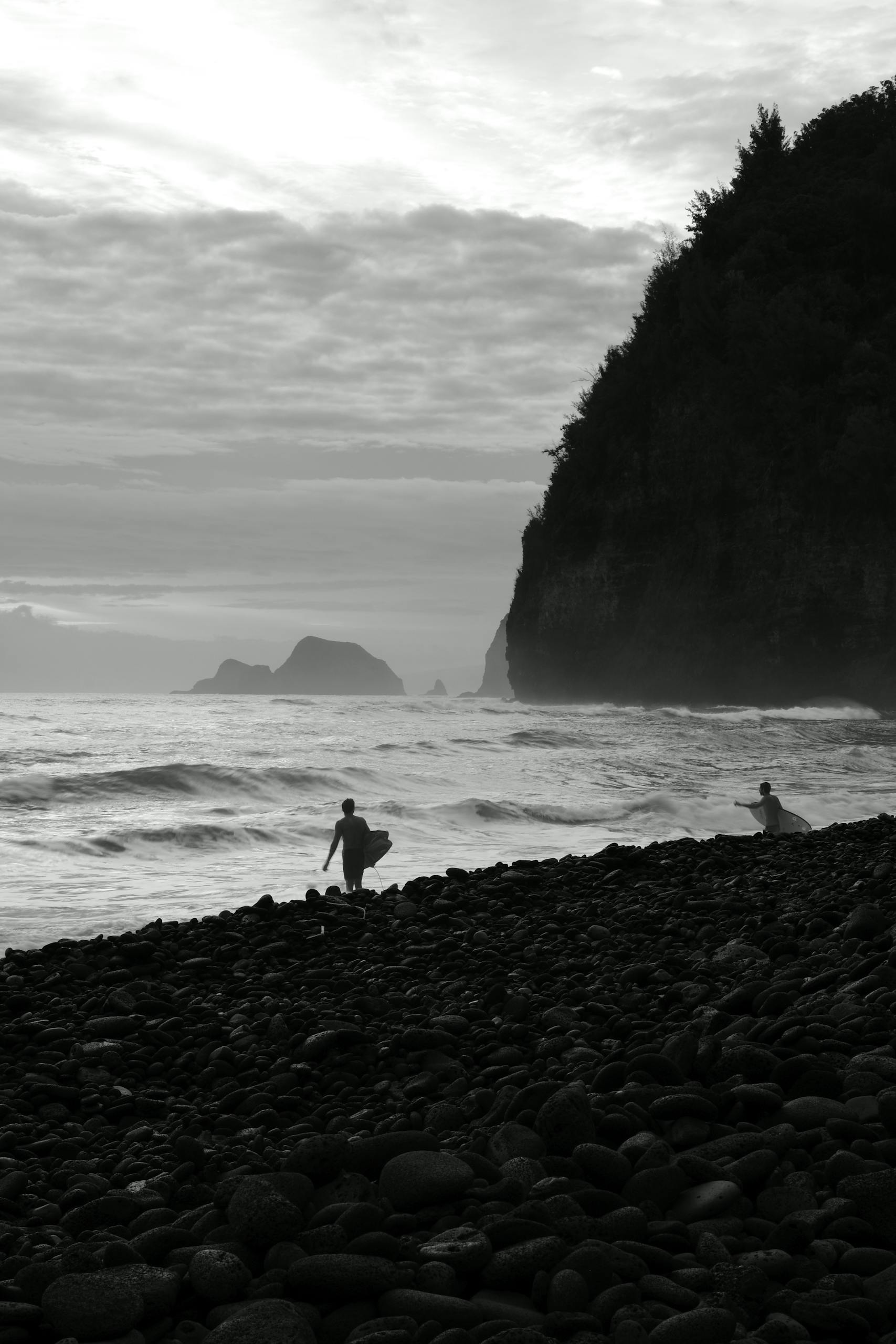 Black and white photo of surfers at a rocky beach in Hawaii backed by a stunning cliff.